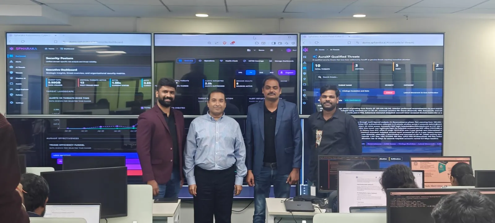Four people on a security operations floor, standing in front of three large screens each running a view of the Spharaka autonomous cyber defence console.