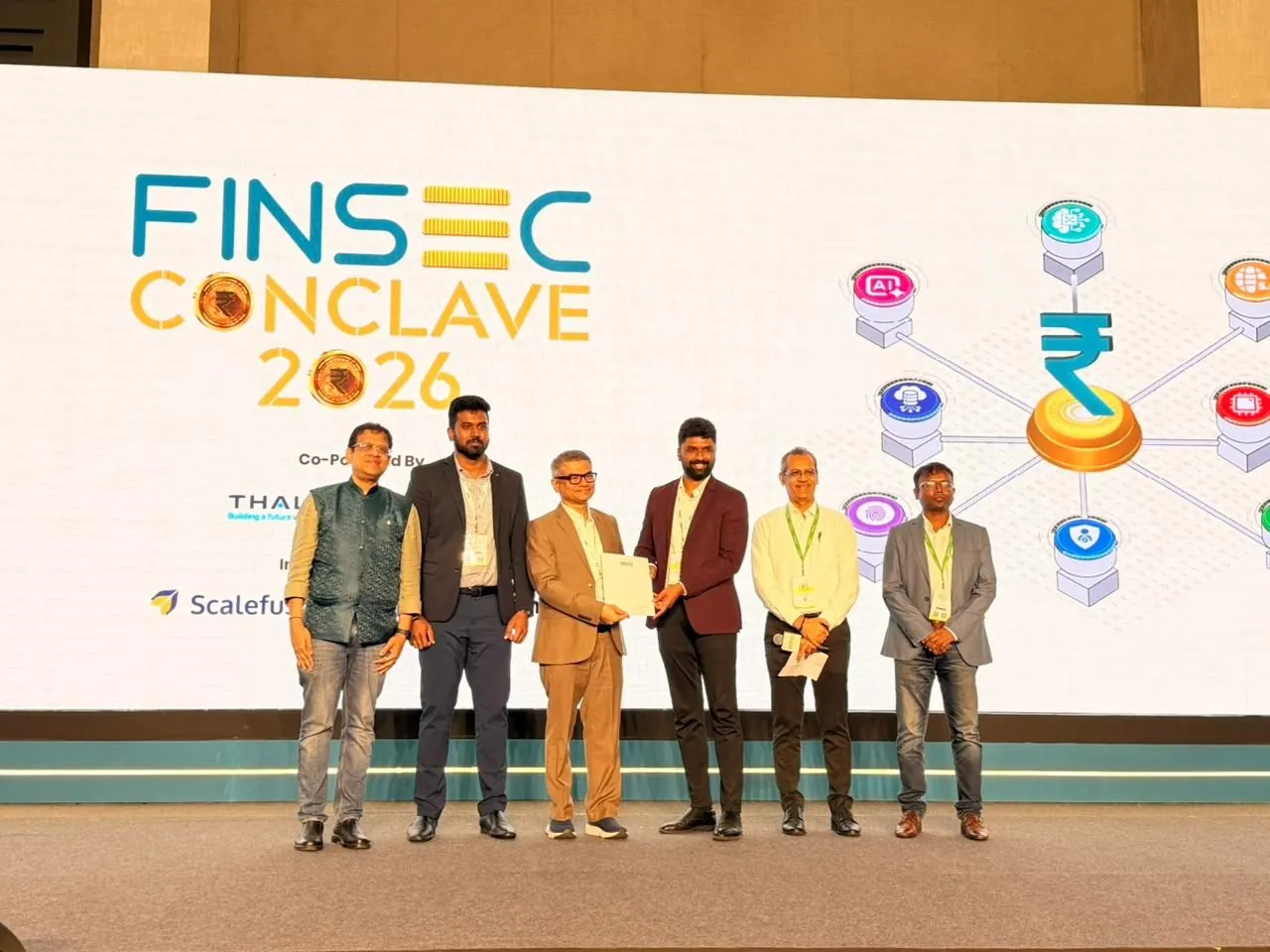 Six people on stage at the DSCI FINSEC Conclave 2026, two of them exchanging a signed document in front of the event backdrop.